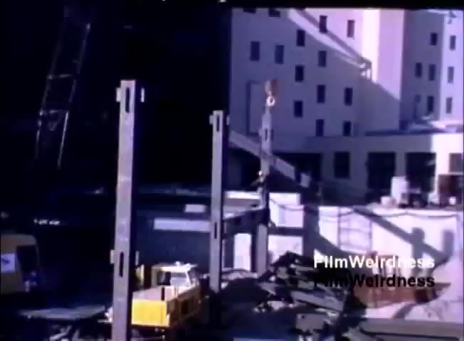 The still depicts a construction site with a focus on a building framework. Steel beams are being erected, suggesting ongoing construction activity. A crane is visible, poised to lift materials, while a small yellow vehicle is stationed nearby. The background features partially completed structures, indicative of an urban development. The visual captures the industrial environment typical of the early 1980s, aligning with the themes of progress and technology in the IBM System 36 sales film.