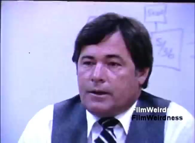 The still depicts a man speaking in a professional setting, likely during a presentation or discussion related to the IBM System 36. He is dressed in business attire with a vest and tie, and appears to be conveying important information. The background features a whiteboard with diagrams or flowcharts, emphasizing a business or technological theme. The overall atmosphere suggests a sales or promotional context from the 1980s.