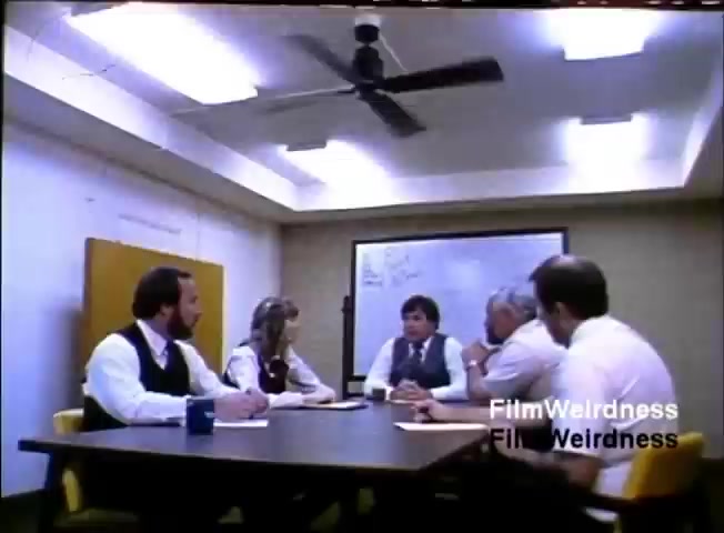 The still features a business meeting set in a small conference room. Five individuals are seated around a rectangular table, engaged in discussion. The atmosphere appears formal, with participants dressed in suits, and a whiteboard is visible in the background. A ceiling fan is overhead, and there are papers and a blue mug on the table, indicating a collaborative environment. The frame captures a moment of interaction, likely centered around the IBM System 36 product being sold during the 1984 presentation.