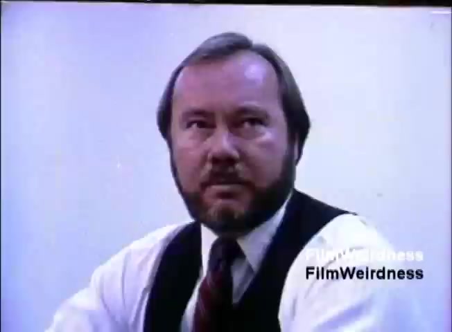 The still from the IBM System 36 sales film features a middle-aged man with a beard, wearing a formal outfit including a vest and tie. He appears to be engaged in discussion, with a serious expression. The background is plain and light-colored, emphasizing his presence in the frame. The image captures a moment likely intended to convey professionalism and authority related to the IBM sales narrative.