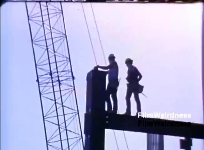 The still shows two silhouetted construction workers standing atop a steel beam at a construction site. A crane is visible in the background, indicating ongoing construction activities. The image captures a clear sky, suggesting a bright day. The focus on the workers emphasizes the human aspect of industrial progress, likely aligning with themes in the sales film about IBM's technology supporting business growth.