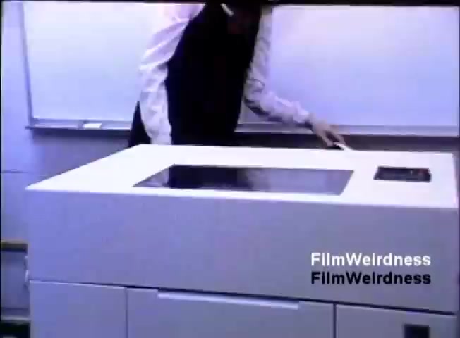 The still shows a person in formal attire interacting with a white IBM System 36 computer. The machine features a flat surface, likely a monitor, and is set in a classroom or office environment with a whiteboard in the background. The person's hand is on the device, indicating demonstration or operation. The setting suggests a sales presentation context from 1984.