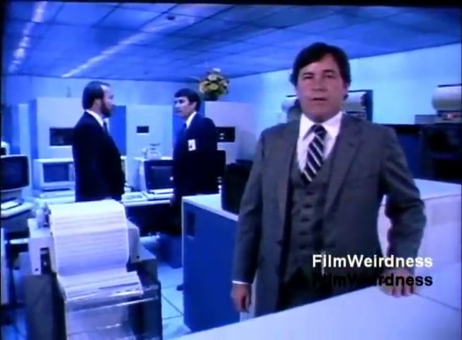 The still features a corporate setting with three men, dressed in formal attire. The foreground shows a man in a suit, possibly addressing the camera or audience. Behind him, two other men engage in conversation, with computers and office equipment visible around them. The environment has a blue tint, typical of the 1980s aesthetic, and suggests a focus on technology, likely emphasizing the IBM System 36's capabilities.
