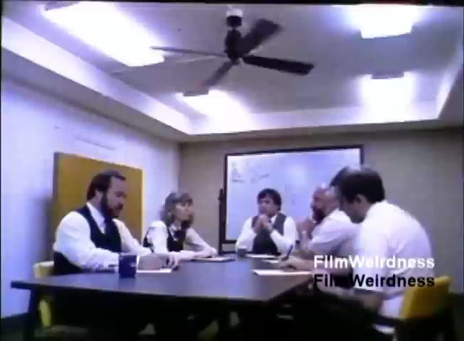 The still from the 'IBM System 36 sales film 1984 - part 1' depicts a business meeting in a conference room. Five individuals, dressed in formal attire, are seated around a table, engaged in discussion. A whiteboard is visible in the background, hinting at a collaborative or strategic dialogue. The overall atmosphere suggests a corporate environment focused on sales or product discussion.