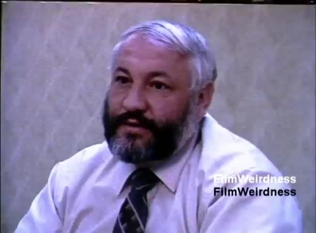 The still depicts a man with a beard, seated and facing the camera. He is wearing a light-colored shirt and a tie, appearing engaged in conversation. The background features a patterned wall, typical of an office or conference setting. This image is part of a sales film promoting the IBM System 36 from 1984, likely intended to illustrate a personal or professional pitch about the product.