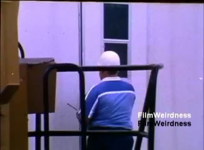 The still depicts a worker wearing a white hard hat and a blue-and-white striped shirt, seated on a platform or lift while focused on a task. The background features a building or structure with a window. The overall tone reflects a work environment, likely related to the context of the IBM System 36 sales film from 1984, highlighting operational aspects of the technology or its setup.