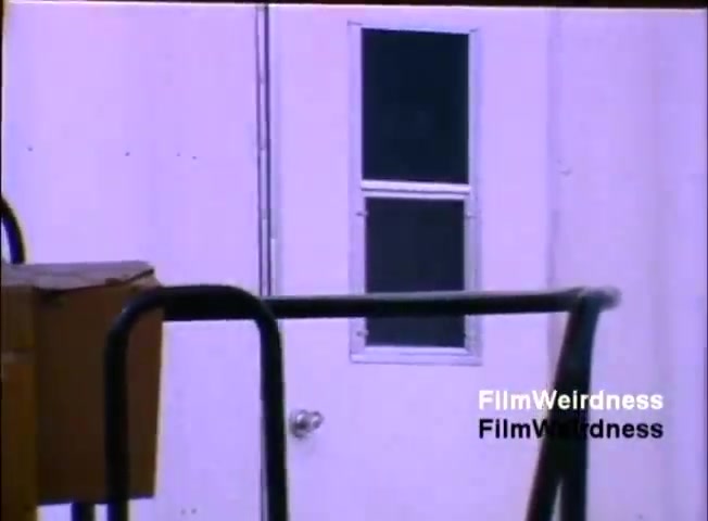 The still from the 'IBM System 36 sales film 1984 - part 1' features a close-up view of a white door with a window, framed by a structure that appears to be part of a larger industrial setting. The door has a simple design, with a knob visible. Portions of equipment or boxes are seen in the foreground, suggesting an environment related to business or technology. The overall mood is utilitarian, reflecting the era's focus on practical solutions in computing.
