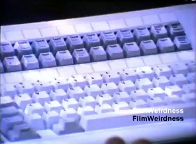 The still from the IBM System 36 sales film showcases a close-up of a vintage computer keyboard. The keys are predominantly white, featuring a simplistic layout typical of the era, likely designed for business users. The lighting gives the keyboard a slight blue hue, emphasizing its modernity for the 1980s. This visual highlights the technological advancement and user-friendly design promoted in the sales film.