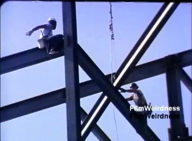 The still from the 1984 IBM System 36 sales film depicts two workers atop a steel structure. One worker is seated on a beam with a bucket, while the other is standing, appearing to handle equipment or tools. The setting is outdoors under a clear blue sky, emphasizing the construction context. The focus highlights industrial labor and teamwork.