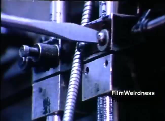The still features a close-up of machinery, likely part of the IBM System 36. It shows metal components, including a lever or blade positioned near a circular part, suggesting a mechanical operation. The focus is on the intricate details of the machinery, emphasizing the engineering involved in the IBM System 36. The color tone appears somewhat bluish, typical of certain film recordings from that era.