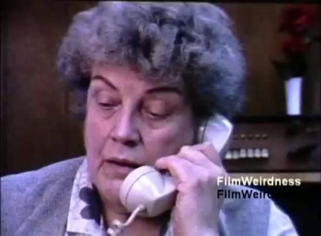 The still features a middle-aged woman with curly hair speaking on a telephone. She appears focused and possibly concerned, engaging in an important conversation. The background is somewhat blurred, suggesting a simple office or home setting, with a hint of plants. The image captures a moment of communication, likely related to the IBM System 36's sales or functionality.