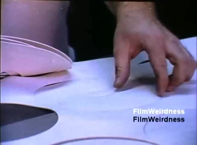 The still shows a close-up of a hand placing or adjusting a paper or material sheet on a surface. The background features circular shapes, suggesting an assembly or production context. This scene likely emphasizes the tactile aspect of the work involved in presenting or utilizing IBM's System 36 technology. The overall tone is practical and industrial, reflecting processes relevant to the sales film's purpose in showcasing the product.