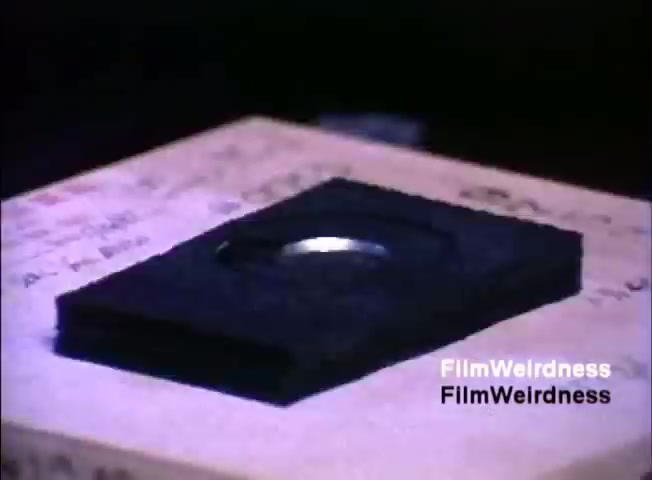 The still features a black, rectangular object with a shiny circular element on top, resting on a surface that has handwritten notes or markings. This setup likely showcases a component or accessory related to the IBM System 36, emphasized in a sales context from the 1984 film. The focus on the object suggests its importance in the system's functionality or presentation.
