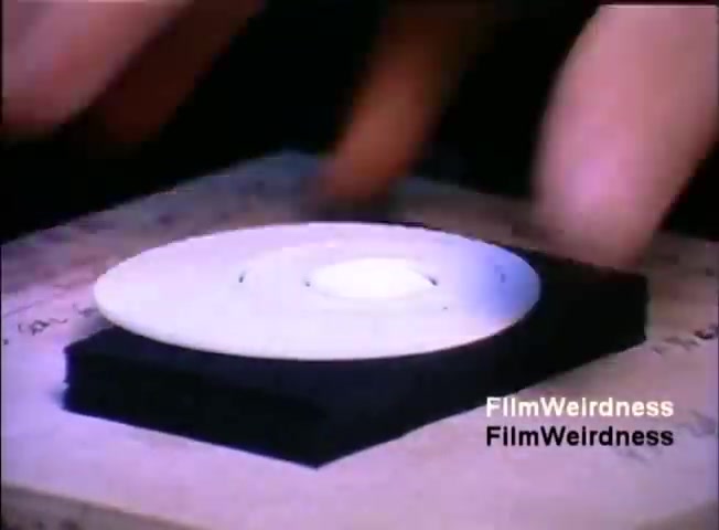 The still features a close-up of a hand interacting with what appears to be a round, white disc placed on a black surface. The disc has a central hole and is set against a wooden background. The context suggests this might be an illustration of technology or a product demonstration linked to the IBM System 36, likely highlighting its components or features in the sales film.