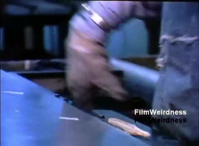 The still from the 'IBM System 36 sales film 1984 - part 1' shows a close-up of a worker's hand, wearing gloves, engaged in a task. The focus is on the hand manipulating an object on a metallic surface, possibly part of the assembly or maintenance of the IBM System 36. The background is blurred, emphasizing the action taking place, and the overall tone suggests a hands-on, technical environment related to computing technology.