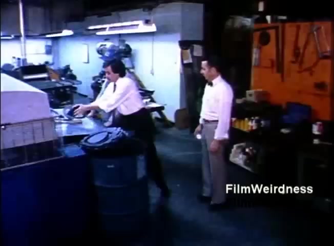 The still depicts two men in a workshop setting, with one dressed in a white shirt and bow tie actively working at a table with equipment, while the other, also in a white shirt, stands nearby observing. The environment appears industrial, featuring equipment and containers, reflecting a business context. The film is part of a sales presentation for the IBM System 36 from 1984, suggesting a focus on demonstrating business efficiency or technology solutions.