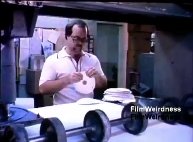 The still features a man wearing glasses and a short-sleeved shirt, working in a somewhat industrial environment. He is holding a circular disc while examining it, likely in relation to the IBM System 36 operations. The background shows machinery and documents, suggesting a workspace focused on production or data handling. The overall atmosphere reflects a mid-1980s setting related to technology or office automation.
