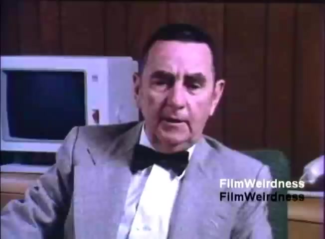 The still features a middle-aged man wearing a suit and bow tie, sitting in an office setting. He appears to be speaking directly to the camera, possibly delivering a sales pitch or explaining features related to the IBM System 36. In the background, an older computer monitor is visible, indicating the time period of the film (1984). The overall ambiance suggests a formal, professional presentation aimed at promoting technology.
