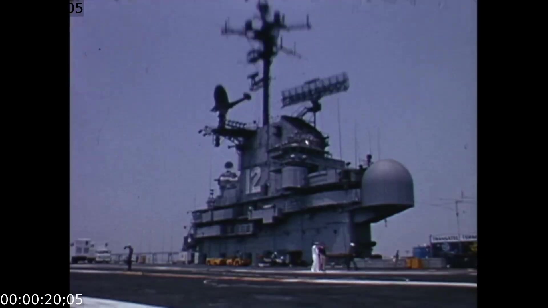 The film still features a large, industrial mobile color TV earth station prominently displayed on a naval vessel, with antennas and equipment atop the structure under a clear blue sky. The camera angle captures the ship's impressive scale as it looms over figures in white suits moving on the deck, highlighting the technological advancements of the time.