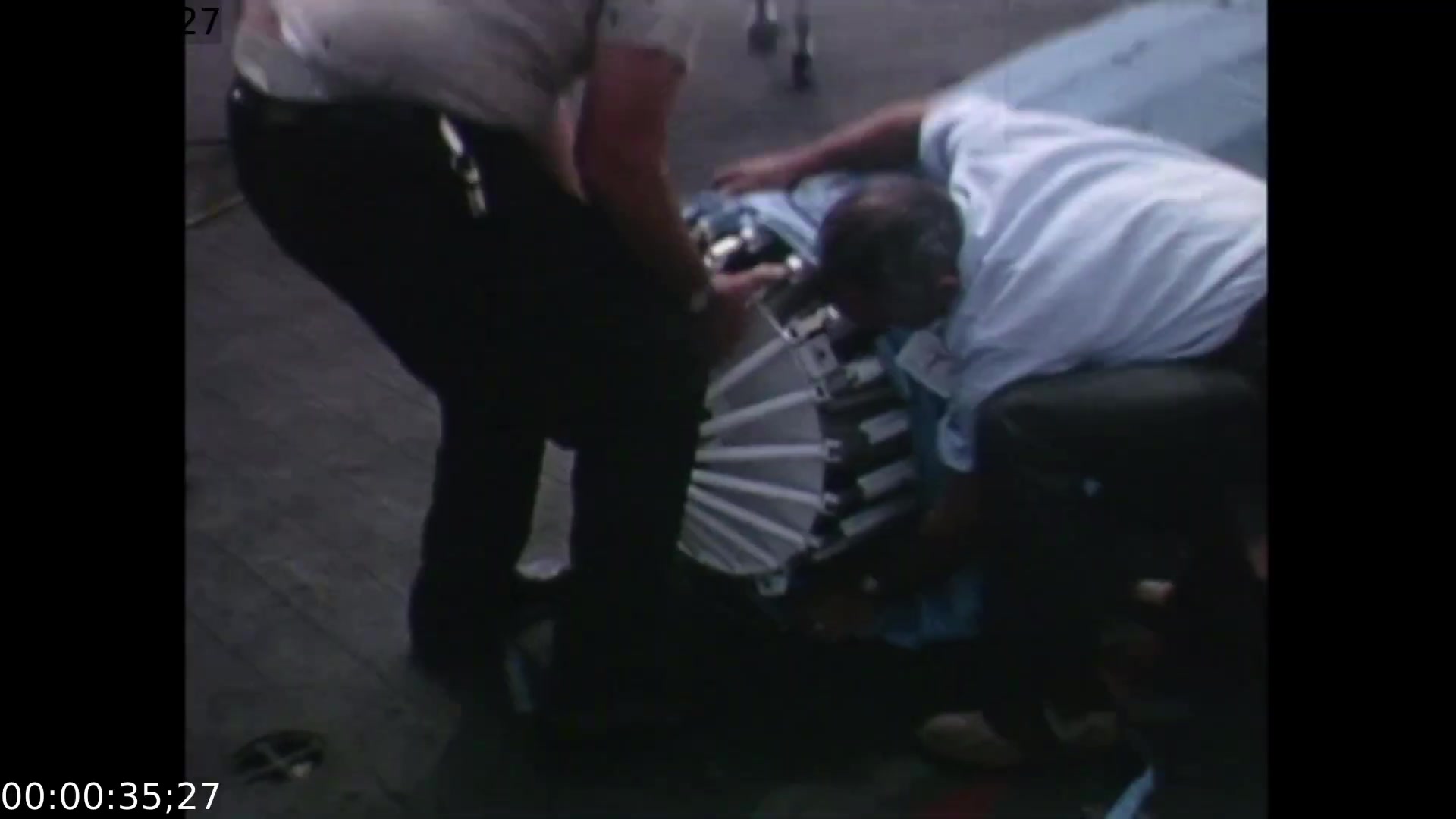 The film still depicts two men bent over a piece of equipment on a deck, suggesting an intense technical setup or repair. The close-up camera angle highlights their focused interactions with the machinery as they prepare for the live telecast of the Apollo 11 splashdown, emphasizing the urgency and collaborative effort behind the broadcast.