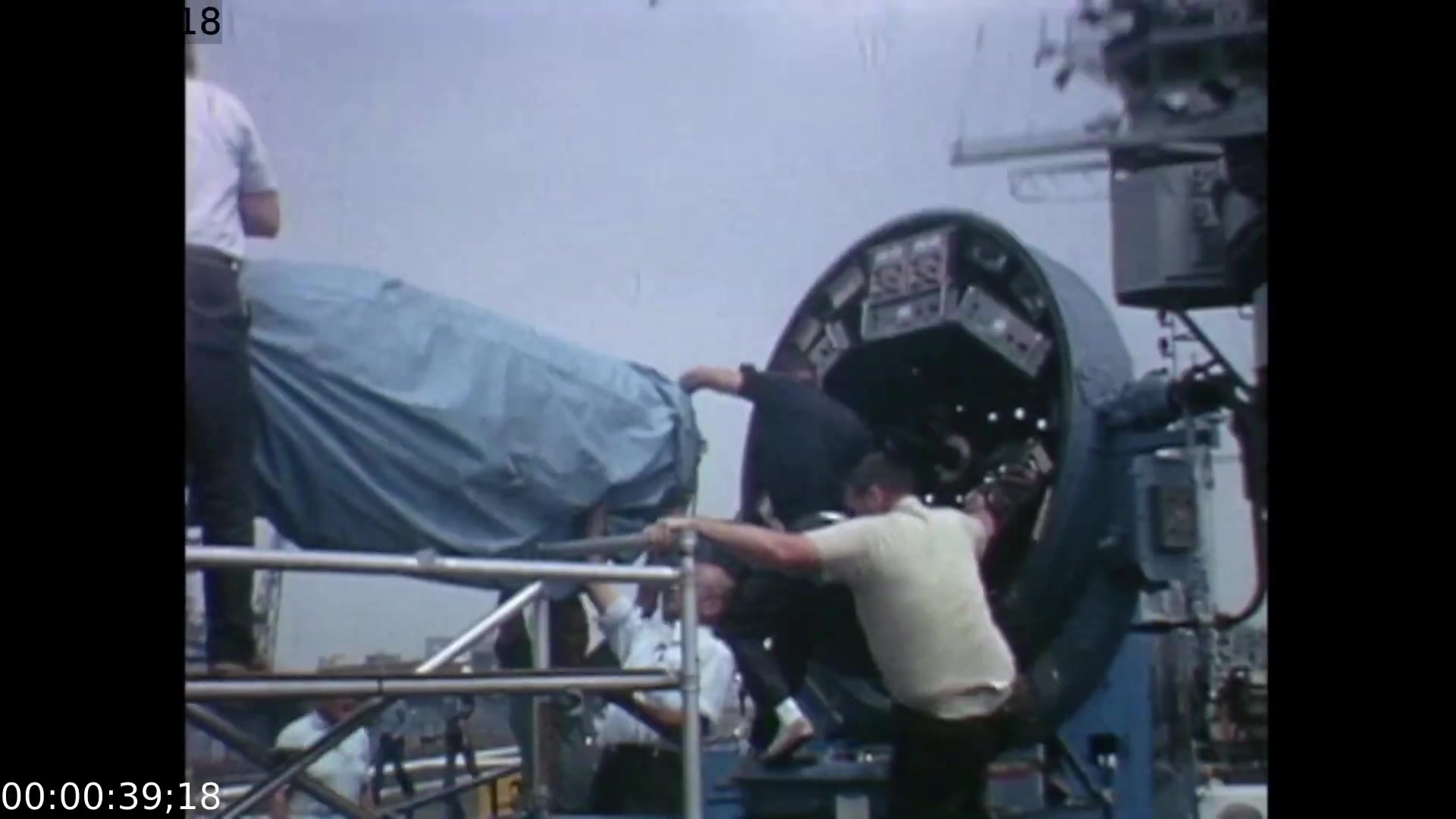 The film still features technicians working on a large, covered piece of equipment, likely a mobile color TV transmission unit, against a backdrop of a cloudy sky. The camera angle captures the dynamic action of crew members as they focus on assembling the gear needed to facilitate live telecasts of the Apollo 11 splashdown and recovery on July 24, 1969.