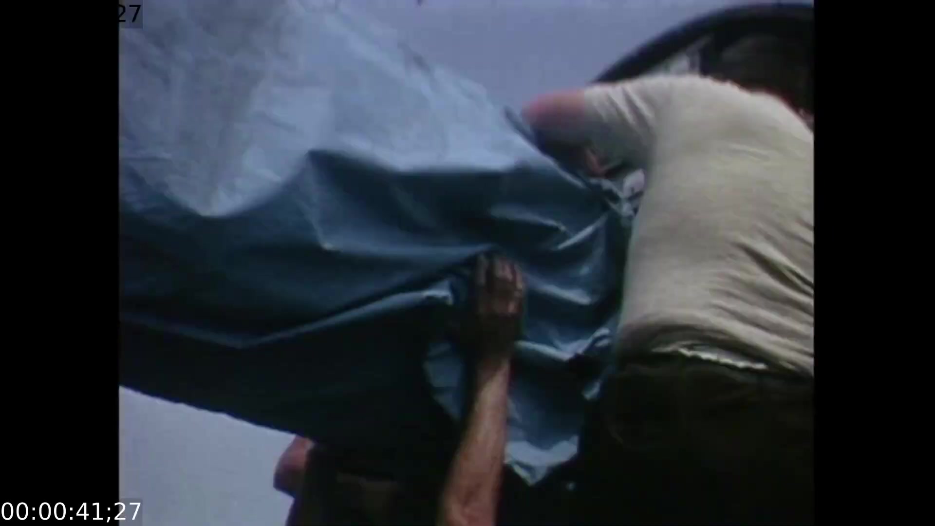The film still showcases a group of technicians focused on assembling equipment, with hands and upper bodies prominently featured as they maneuver a large blue tarp or cover. The camera angle is slightly elevated, capturing the urgency and collaborative effort involved in setting up the mobile color TV earth station for the Apollo 11 splashdown coverage.