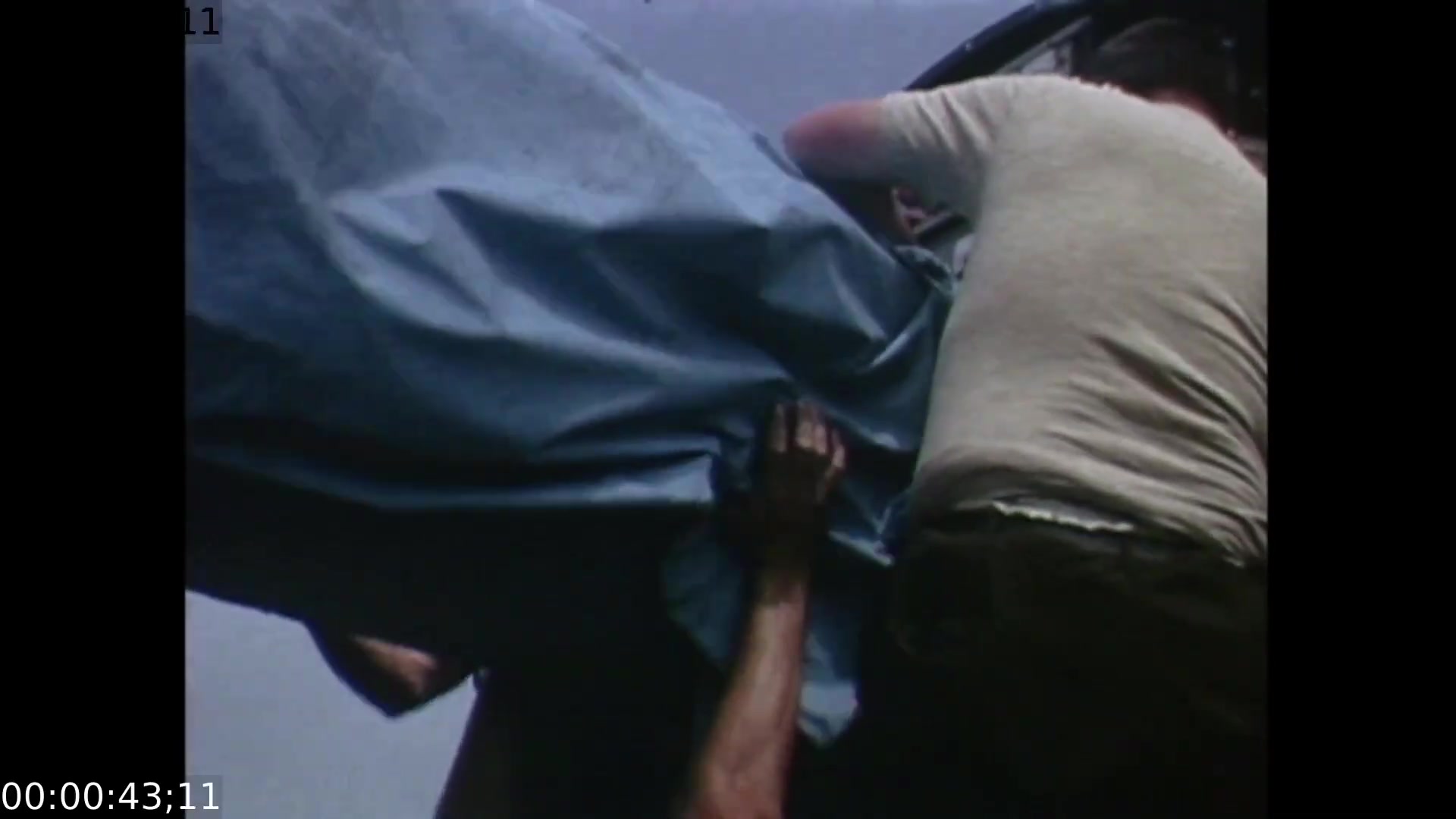 The film still features two individuals engaged in the process of securing a large, blue tarp, likely related to the Apollo 11 splashdown event. The camera angle is close-up, focusing on the hands and physical effort involved, emphasizing the urgency and teamwork required in this moment.