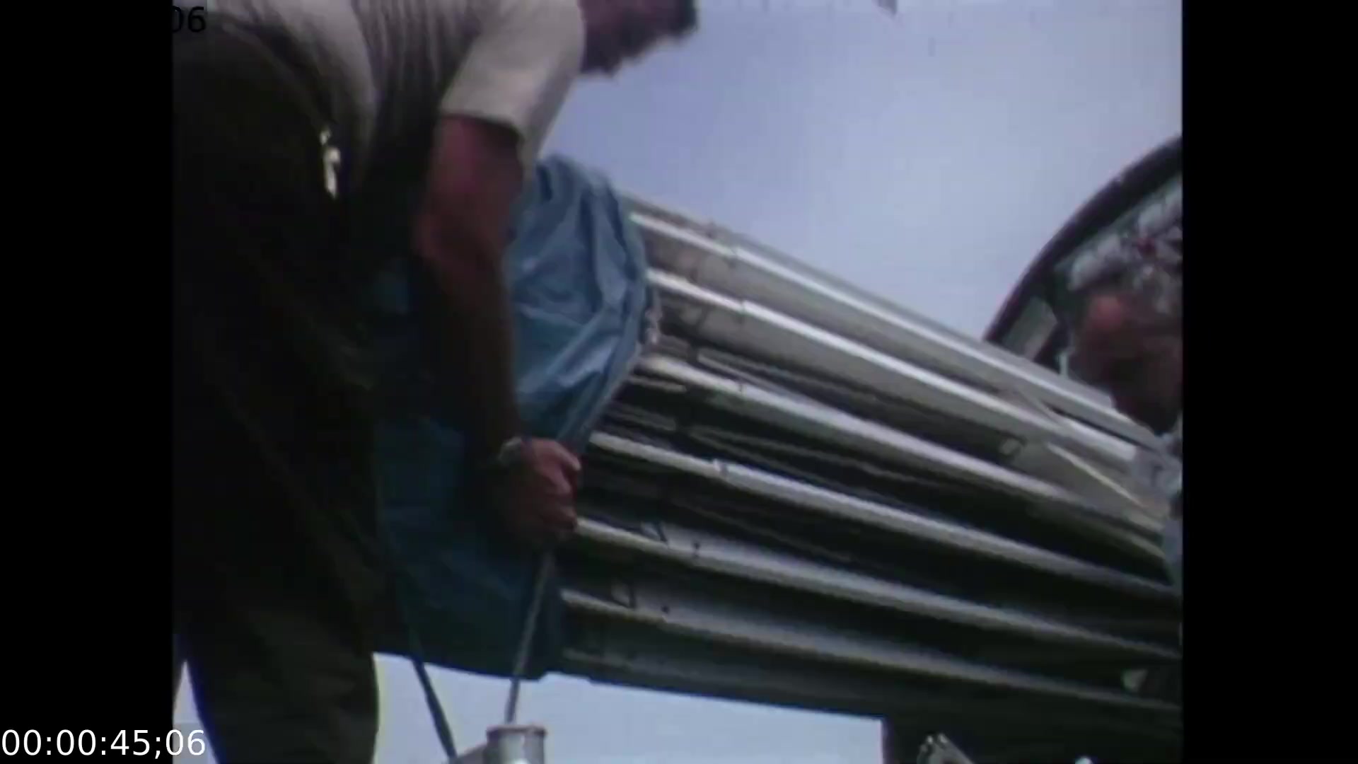 The film still shows two technicians in the process of assembling equipment for the mobile color TV earth station, focused on a large, folded contraption covered with a blue tarp. The camera angle is positioned low, capturing the workers from a slight upward perspective against a backdrop of a partly cloudy sky, emphasizing their task and the scale of the equipment.