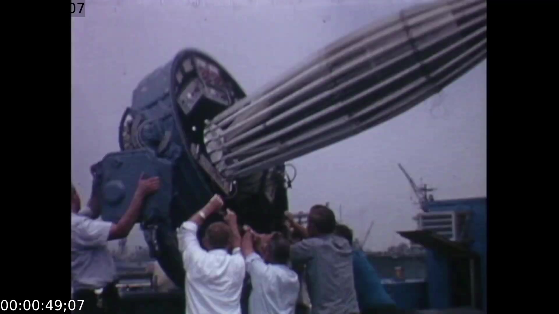The film still captures a group of technicians collaborating to set up the large, intricately designed mobile color TV earth station against an industrial backdrop, likely managing equipment essential for broadcasting. The camera angle emphasizes their teamwork and the scale of the technology involved in transmitting the historic Apollo 11 splashdown.