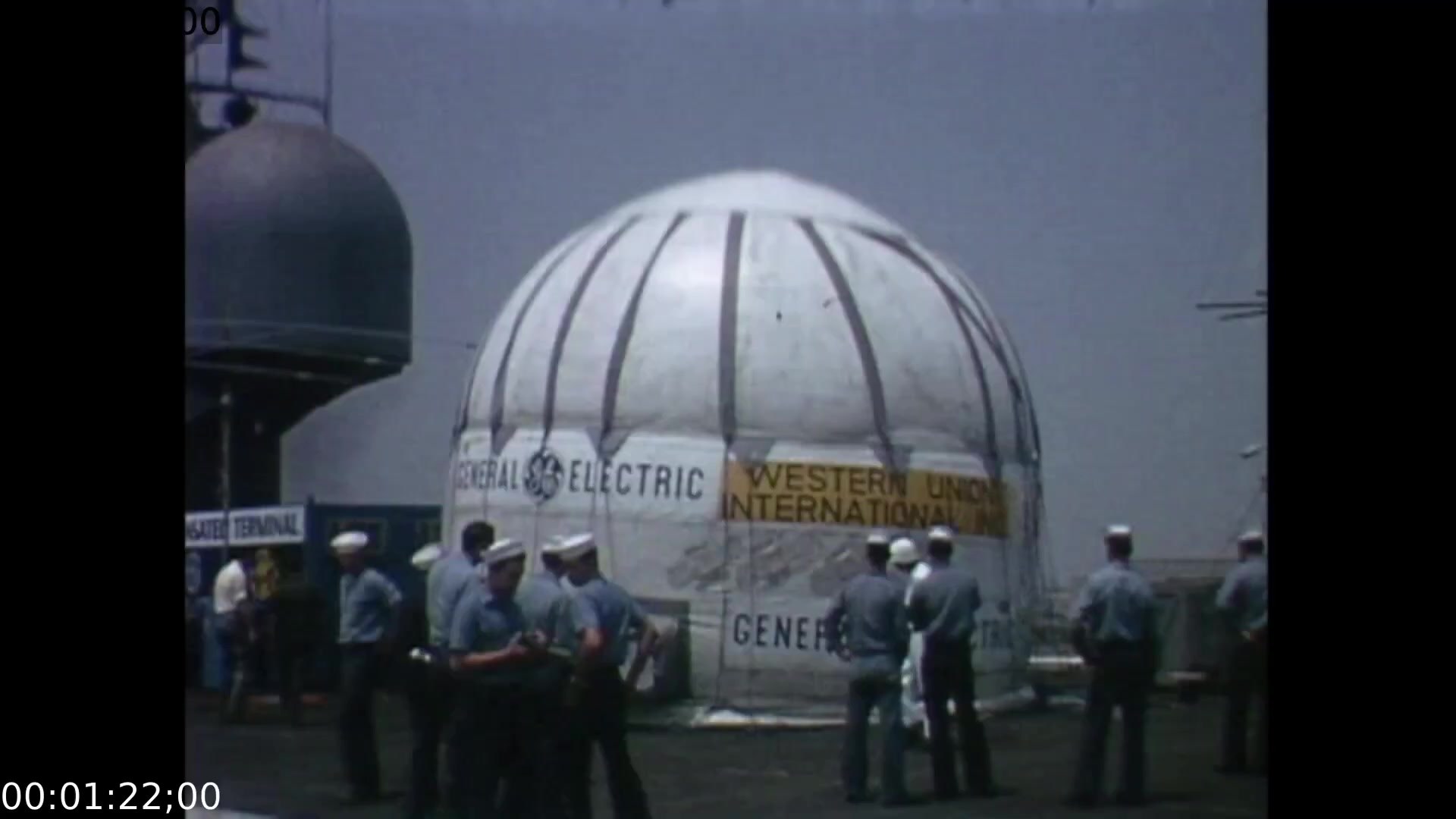 The still captures a group of personnel, likely from the mission or media crew, gathered in front of a large, dome-shaped structure branded with 'Western Union International' and 'General Electric.' The camera angle appears to be positioned at eye level, showcasing the collaboration between the two companies and the atmosphere surrounding the historic Apollo 11 splashdown event.