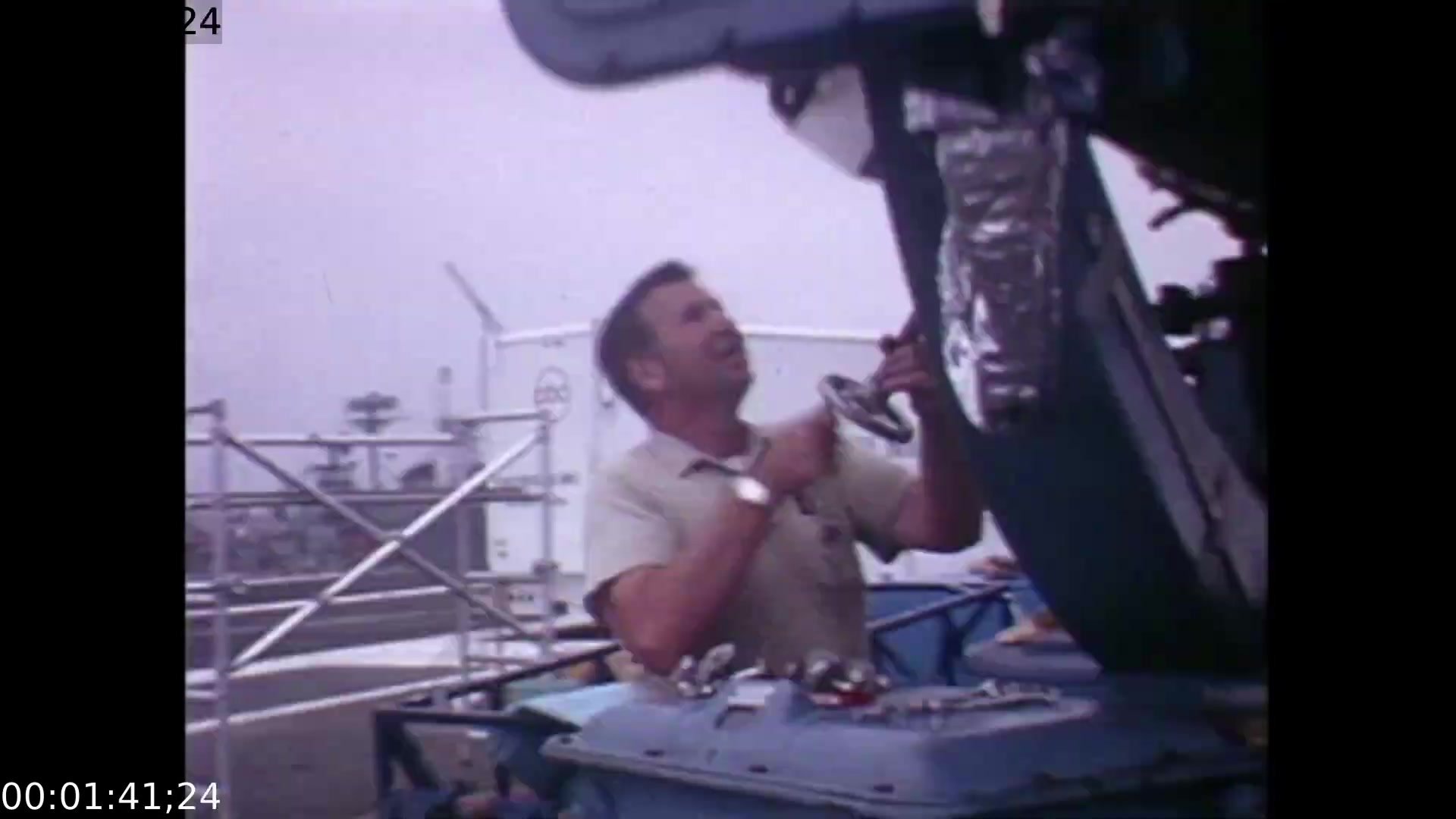 The still features a technician working intently on equipment at a mobile color TV earth station during the Apollo 11 mission. Shot from a low angle, it emphasizes the man's focused expression and highlights the machinery and surrounding scaffolding, capturing the atmosphere of anticipation and technical expertise as the team prepares for the live telecast.