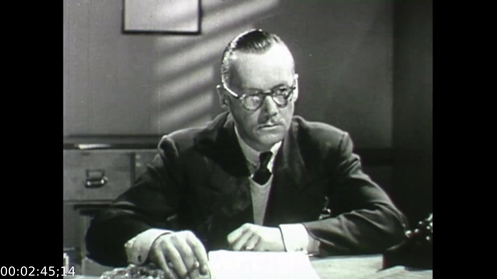 The still from 'Feelings of Depression' (1950) features a somber-looking man, identified as John Murray, sitting at a desk in a dimly lit room. He is dressed in a formal suit and tie, wearing glasses, and appears contemplative or troubled as he gazes down at the document in front of him. The scene captures a moment of introspection, reflecting the film's exploration of mental health struggles and the impact of depression on everyday life. The use of lighting and modest office setting underscores the seriousness of the topic.