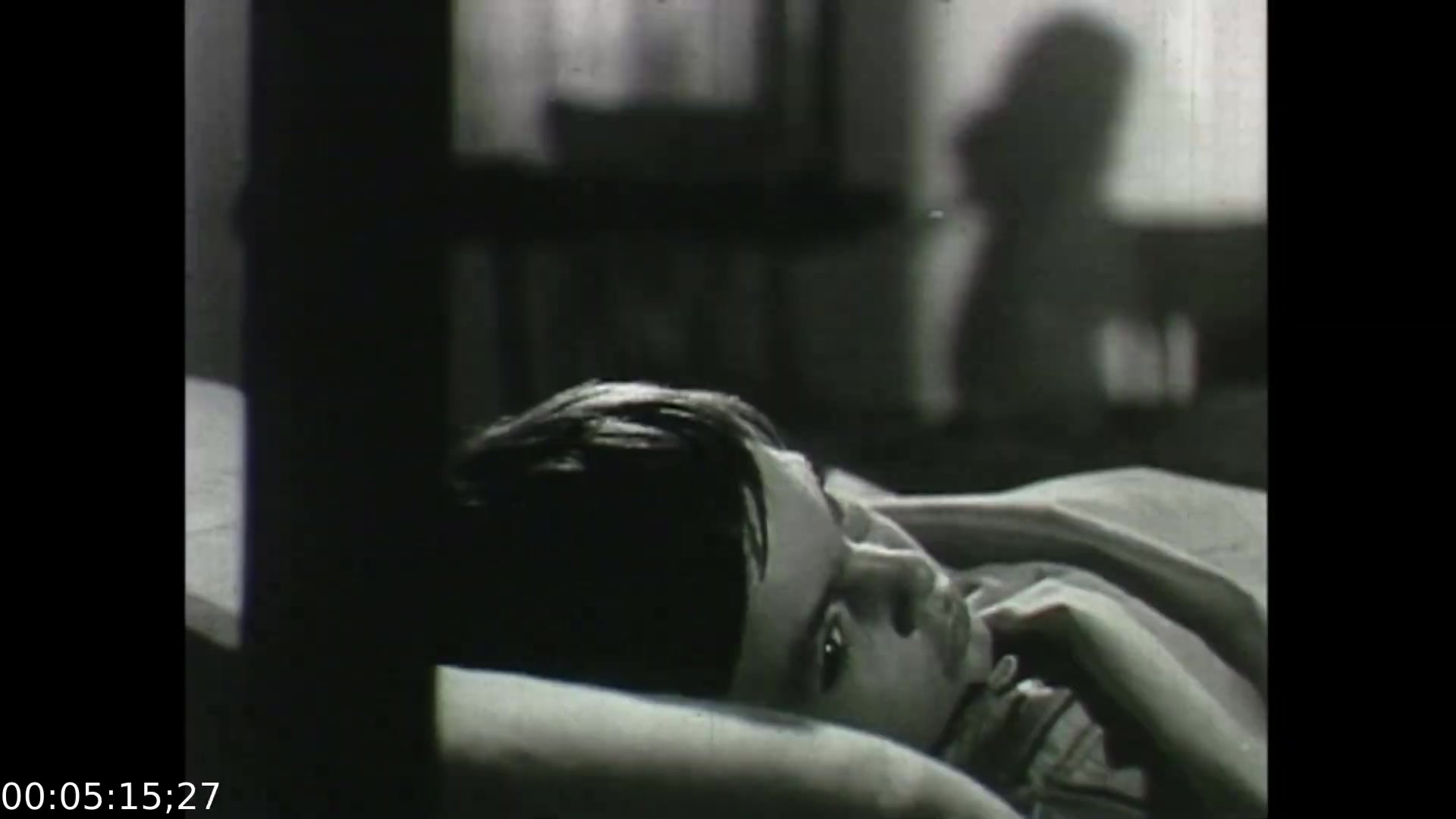 The still from 'Feelings of Depression' features a young boy lying in bed, seemingly deep in thought or distress. The lighting and stark black-and-white imagery enhance the sense of somber contemplation. This moment likely illustrates the internal struggle associated with depression, reflecting themes of emotional turmoil and isolation. The film addresses the impact of mental health on individuals and their families, set against a historical backdrop of limited understanding and resources for mental health issues in the 1950s.