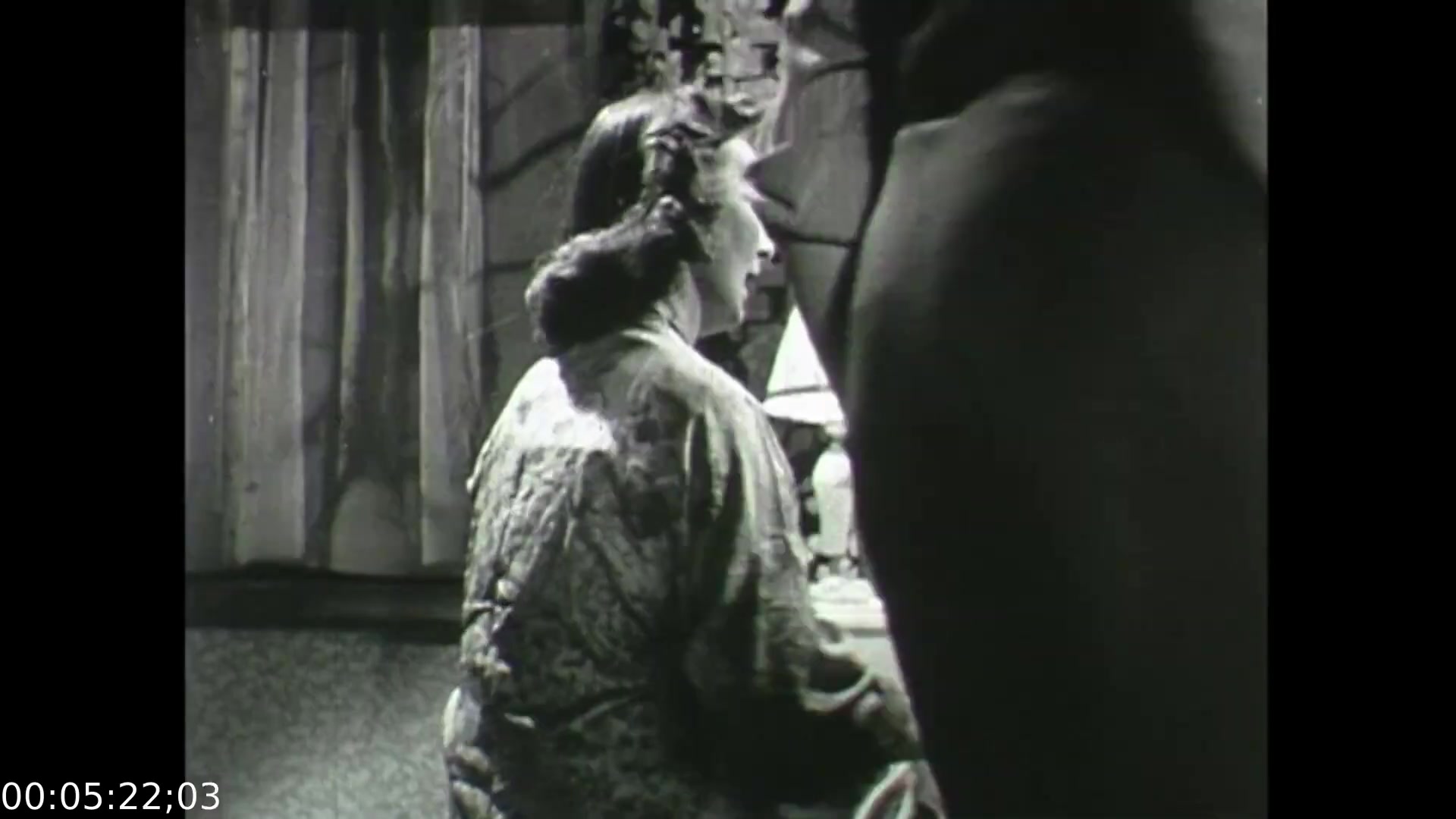 The still from 'Feelings of Depression' depicts a woman turned slightly away from the camera, sitting at a desk with a lamp. The setting has a somber atmosphere, emphasizing the themes of emotional struggle and introspection. Shadows play a significant role, hinting at underlying tension and the gravity of the subject matter. The film aims to educate viewers about the complexities of depression through personal narratives.