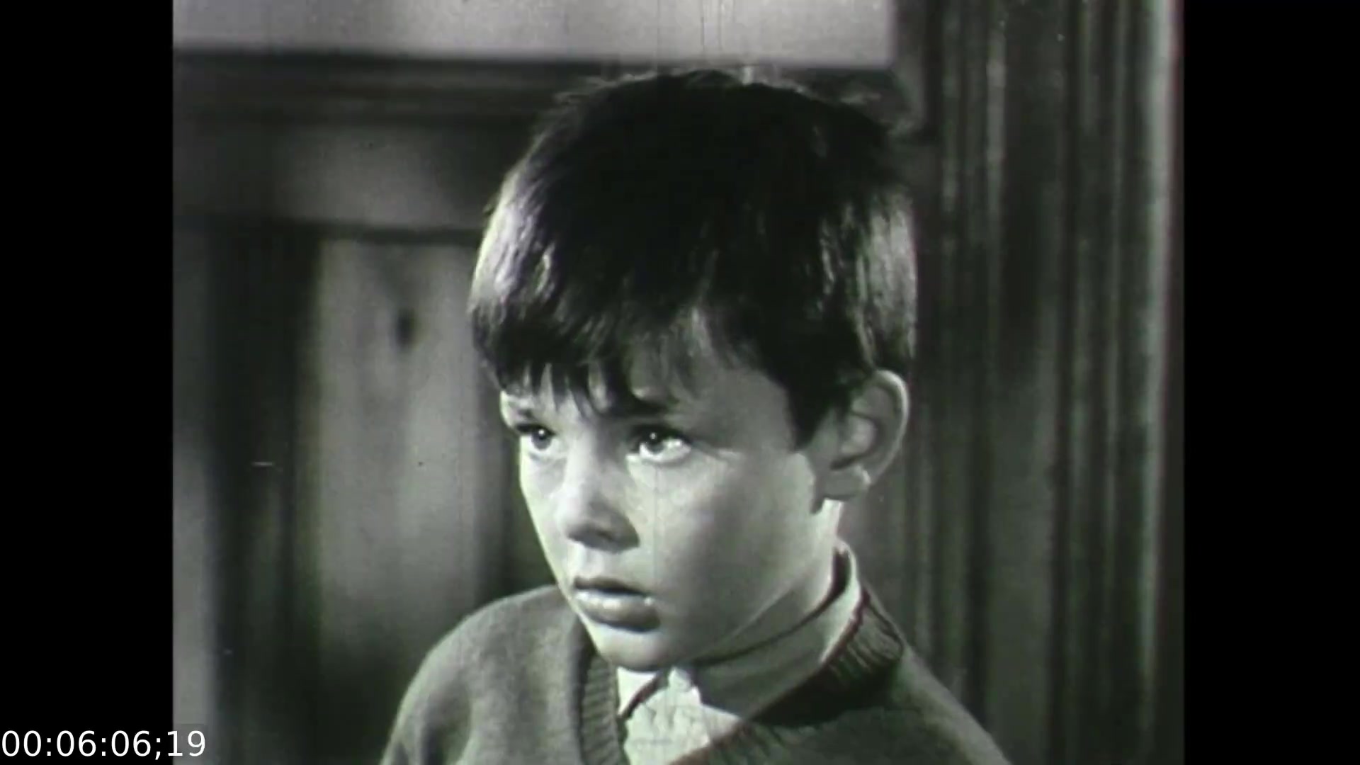 The still from 'Feelings of Depression' (1950) features a young boy with a serious expression, reflecting a sense of concern or confusion. His attire suggests a 1950s setting, and the black-and-white film emphasizes the emotional gravity of the moment. This scene likely illustrates the film's exploration of the impact of mental health struggles on family dynamics, highlighting the boy's emotional response to his surroundings, specifically relating to the challenges faced by his father, John Murray.