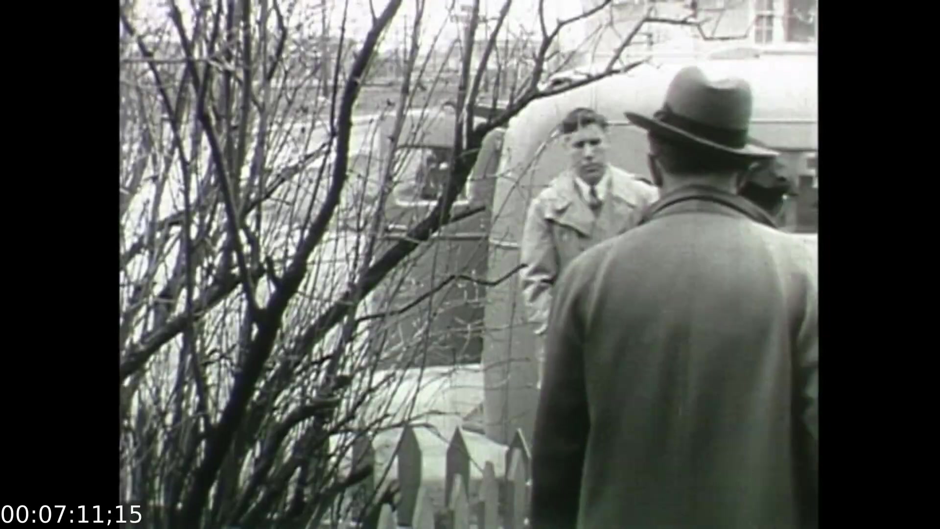 The still from 'Feelings of Depression' depicts a solemn moment between two men, set against a backdrop of bare trees and a picket fence. One man, dressed in a trench coat and hat, faces another in a lighter coat, conveying a sense of tension or concern. The black-and-white aesthetic enhances the film's exploration of emotional struggles, reflecting the somber themes of depression and human connection in post-war society. This scene illustrates the personal interactions within the larger context of mental health issues during the 1950s.