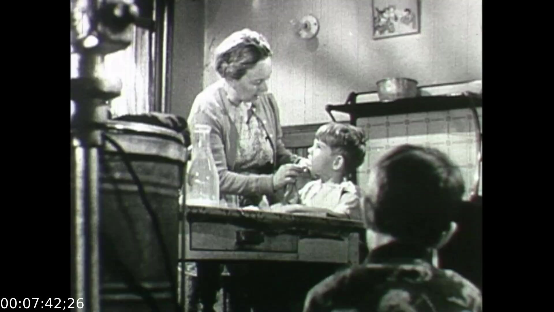 The still from 'Feelings of Depression' (1950) depicts a domestic scene in a kitchen where a woman gently administers care to a young boy, likely conveying warmth and nurturing. This moment reflects themes of family and everyday life, emphasizing the emotional connections that are significant within the context of mental health. The setting suggests a post-war Canadian home, which aligns with the film's exploration of the psychological struggles faced by individuals, like John Murray, and the impact of emotional well-being on family dynamics.