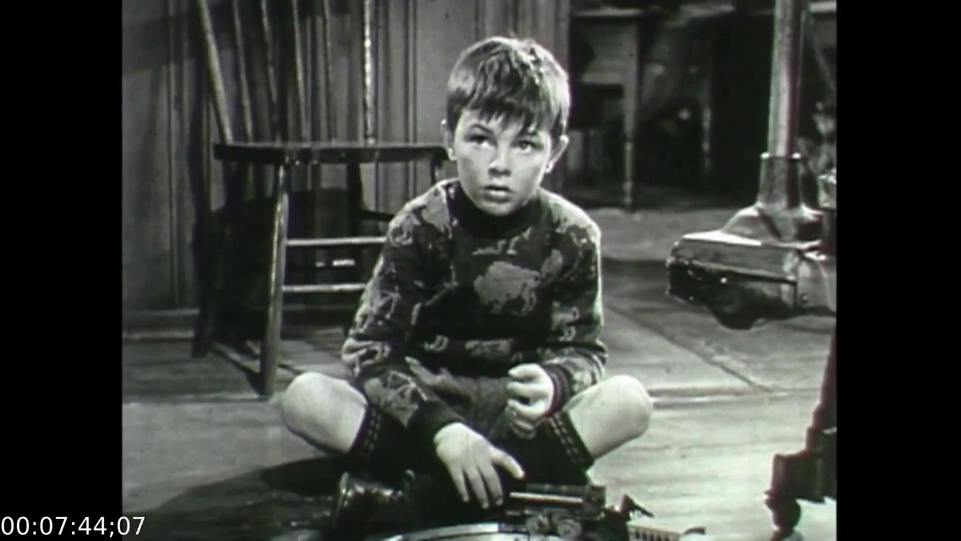 The still from 'Feelings of Depression' depicts a young boy sitting on the floor, clutching a toy gun and looking up with a concerned expression. He is dressed in a patterned sweater and shorts, suggesting a childlike innocence contrasted with his serious demeanor. In the background, a rocking chair and other household items evoke a domestic setting, reinforcing the film's exploration of emotional struggles within a family context. The image captures a moment of vulnerability, reflecting the themes of despondency and the impact of mental health issues.