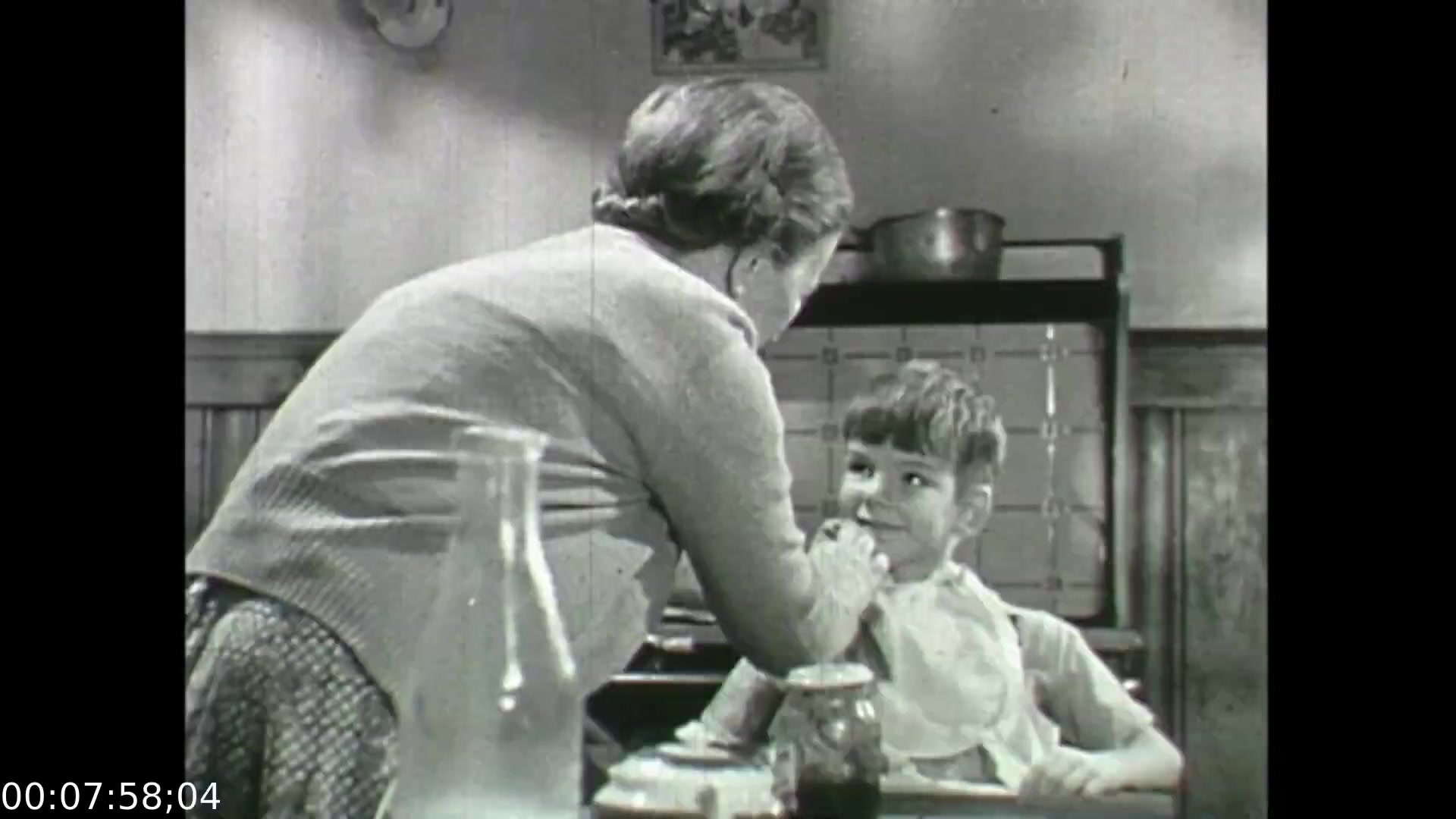 The still from 'Feelings of Depression' (1950) features an intimate moment between an elderly woman and a young boy in a kitchen setting. The woman, leaning toward the boy, appears to be engaging him gently, suggesting a nurturing relationship. The boy, looking up with a slight smile, seems to respond positively. This interaction reflects themes of care and familial support, contrasting with the film's broader exploration of mental health struggles exemplified by the character John Murray. The black-and-white format captures the historical context, emphasizing the societal attitudes toward emotional well-being during that era.
