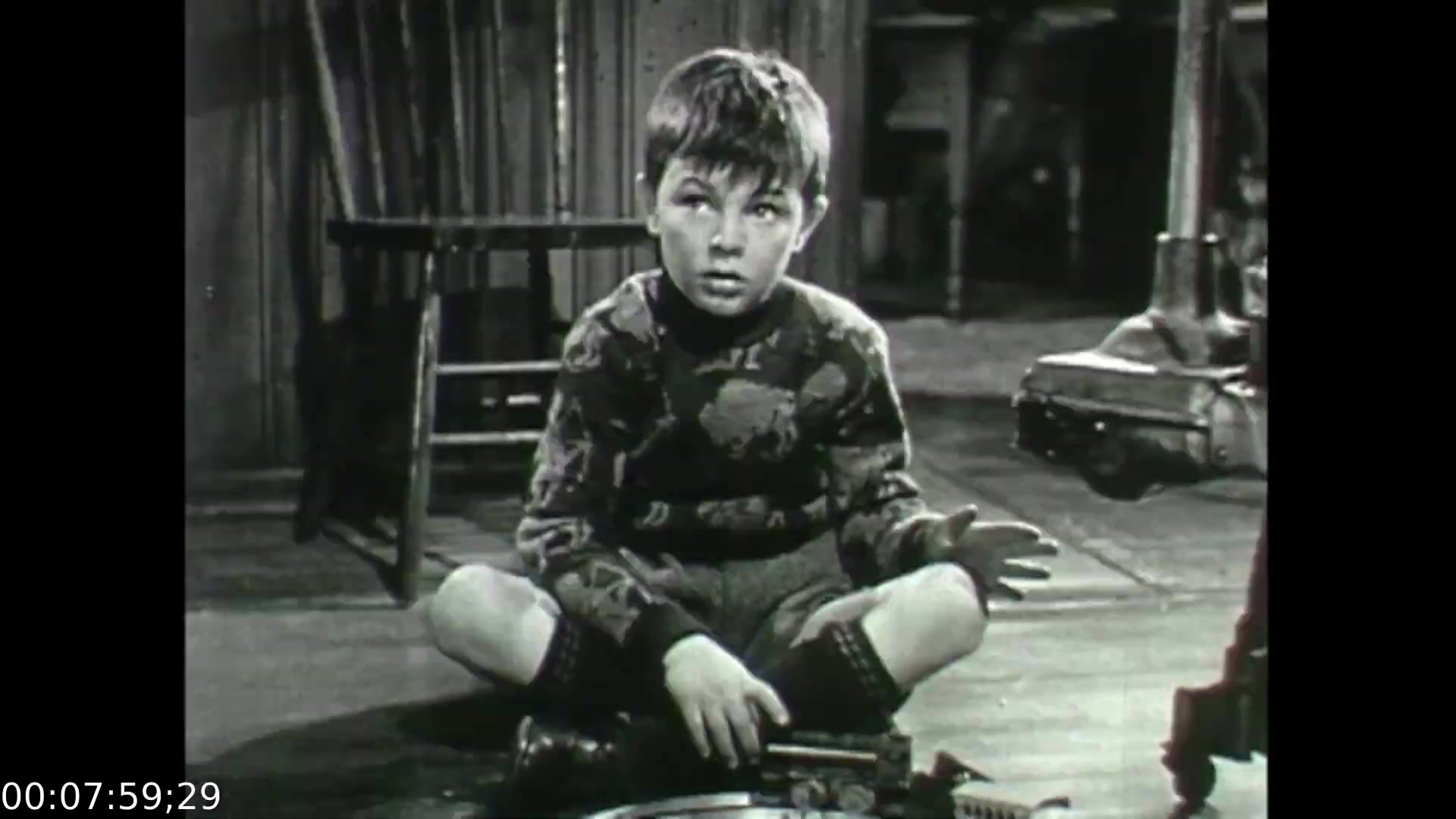 The still features a young boy sitting on the floor, appearing contemplative and engaged in a serious conversation. He is dressed in a patterned sweater and shorts, with a toy or device in his lap. The dimly lit surroundings suggest a homey yet somber atmosphere, complementing the film's exploration of emotional struggles. This scene likely captures a moment reflecting the theme of childhood innocence amidst complex emotions, illustrating the roots of depression as addressed in the film.