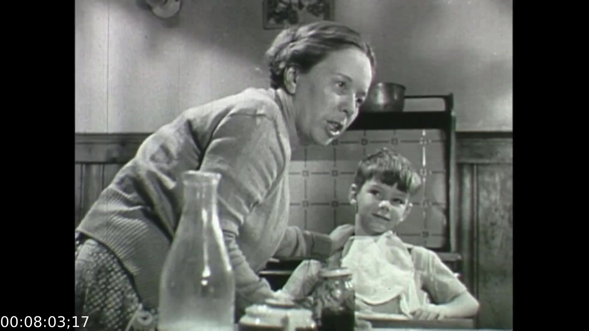 The still from 'Feelings of Depression' (1950) depicts a mother interacting with her young son in a domestic setting. The woman appears engaged, possibly in a conversation or preparing a meal, while the boy, with a slight smile, seems to be enjoying the moment. This scene highlights familial dynamics and the everyday struggles emphasized in the film, which addresses the impact of mental health on family life, particularly through the lens of a father's experiences with depression. The setting adds to the film's exploration of emotional challenges in a historical context.