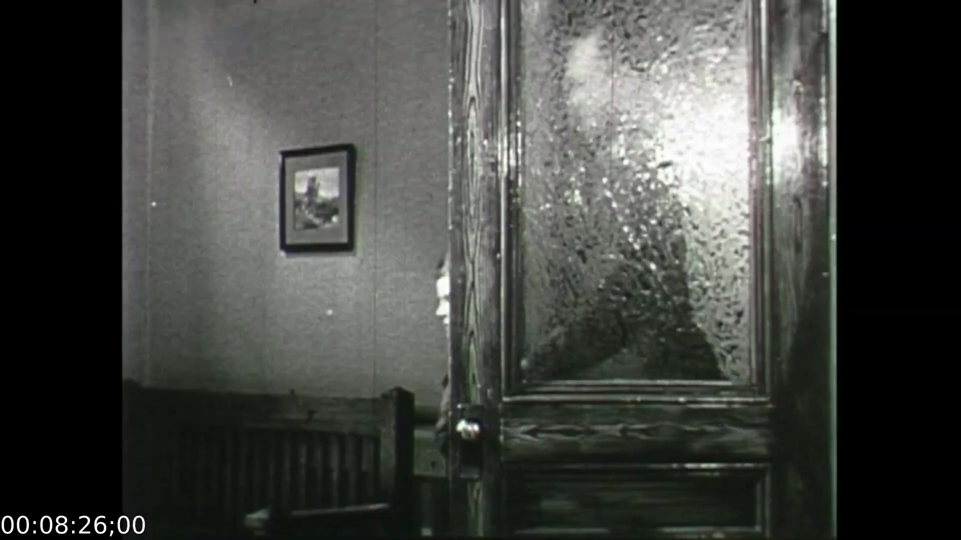 The still from 'Feelings of Depression' portrays a moment of introspection, capturing a glimpse of a door with a textured glass panel. The room appears simple, furnished with a wooden bench and a framed picture on the wall. The focus on the door suggests a threshold between the inner struggles of John Murray and the outside world, symbolizing his battle with despondency. This framing emphasizes isolation and the emotional weight of his experiences, consistent with the film's themes of mental health education in the 1950s.