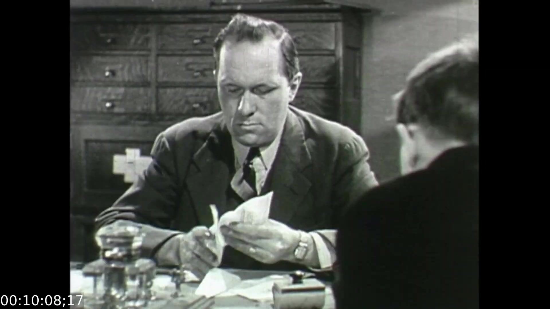The still from 'Feelings of Depression' (1950) depicts a tense moment in a small, somber office setting. The central figure, a businessman, is shown intently focused on a piece of paper, possibly reflecting his inner turmoil and struggles with depression. His furrowed brow and serious demeanor suggest deep contemplation. In the background, a second figure is partially visible, hinting at a conversation or therapeutic interaction. This scene captures the film's exploration of mental health and the challenges faced by individuals like John Murray during that era.