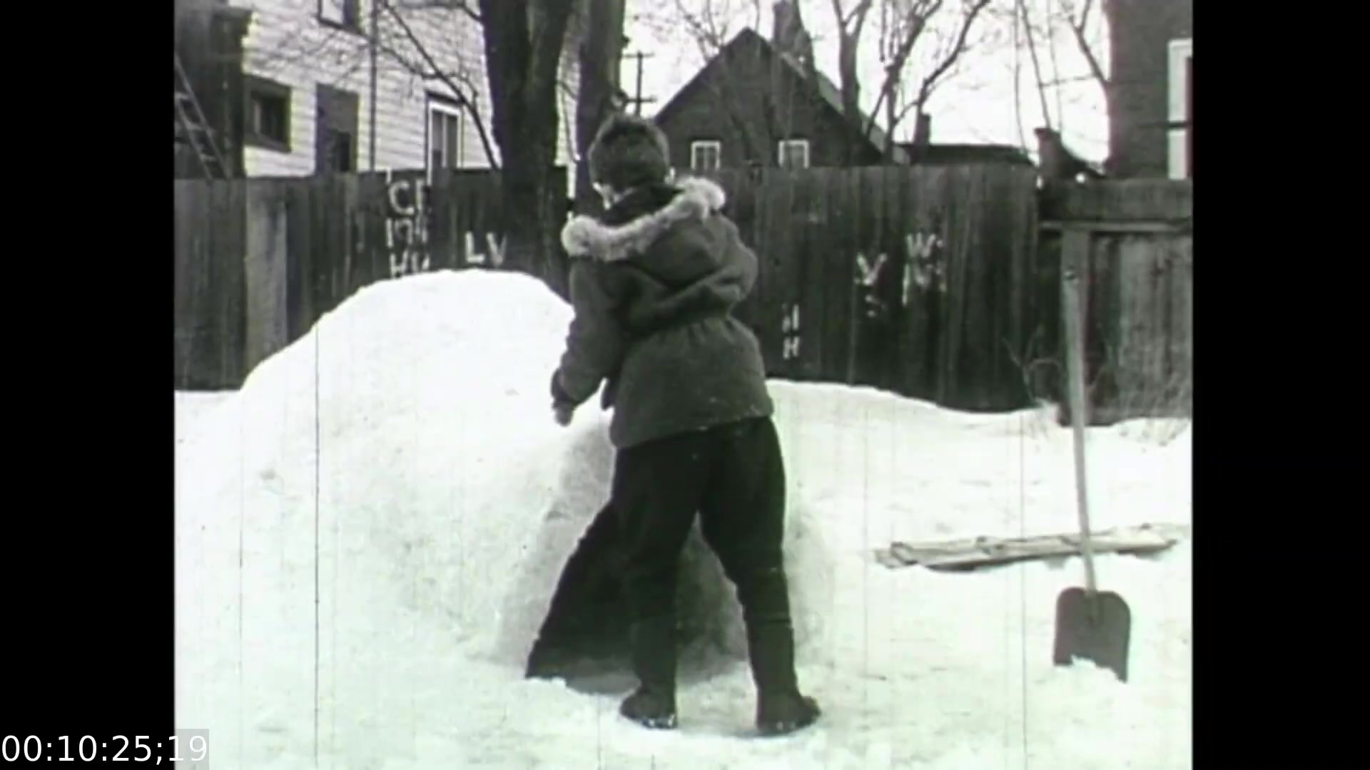 The still from *Feelings of Depression* (1950) depicts a child building an igloo in a snowy backyard. The setting features a wooden fence with visible graffiti and bare trees, suggesting a cold winter environment. The image highlights themes of childhood resilience and playfulness, contrasting against the film's serious exploration of mental health, particularly the struggles faced by the character John Murray. This scene captures a moment of innocence and activity, providing a visual counterpoint to the film's examination of depression.