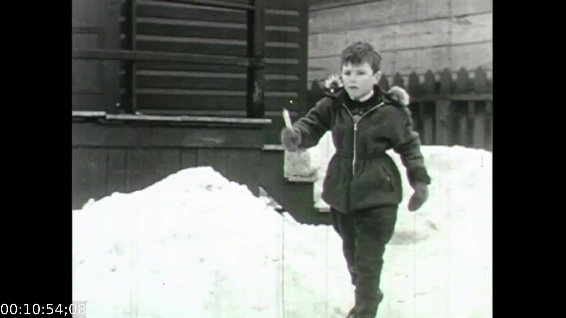 The still from 'Feelings of Depression' (1950) depicts a young boy walking through a snowy environment, wearing a winter jacket and gloves, suggesting a cold climate. He appears to be holding a stick, possibly engaged in play or exploration. The backdrop features a wooden structure, indicative of a modest setting, contributing to the film's exploration of human emotion and everyday life. This scene subtly contrasts with the film's focus on adult struggles, reflecting the innocence of childhood amidst themes of despondency.