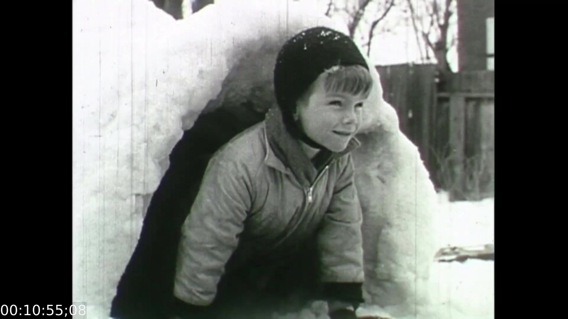 This still from 'Feelings of Depression' shows a young child emerging from a snow fort, smiling. The scene captures a moment of innocence and joy, contrasting the film's serious exploration of mental health issues experienced by adults. This juxtaposition reflects the idea of childhood happiness amidst the complexities of emotions and the struggles depicted in the film. The black and white aesthetic emphasizes the historical context of the 1950s, further highlighting societal perceptions of mental health during that era.
