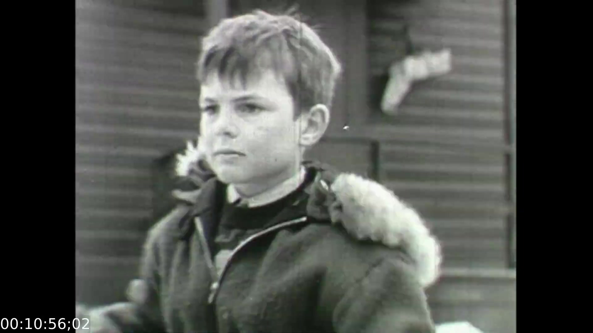 The still from 'Feelings of Depression' (1950) features a young boy wearing a coat with fur lining, gazing pensively toward the camera. The image captures a moment of introspection, reflecting the emotional struggles portrayed in the film. Set against a backdrop of wooden structures, the scene symbolizes the early experiences of despondency that are central to the narrative, illustrating how such feelings can impact individuals from a young age. The film serves as an educational dramatization aimed at raising awareness about mental health issues.