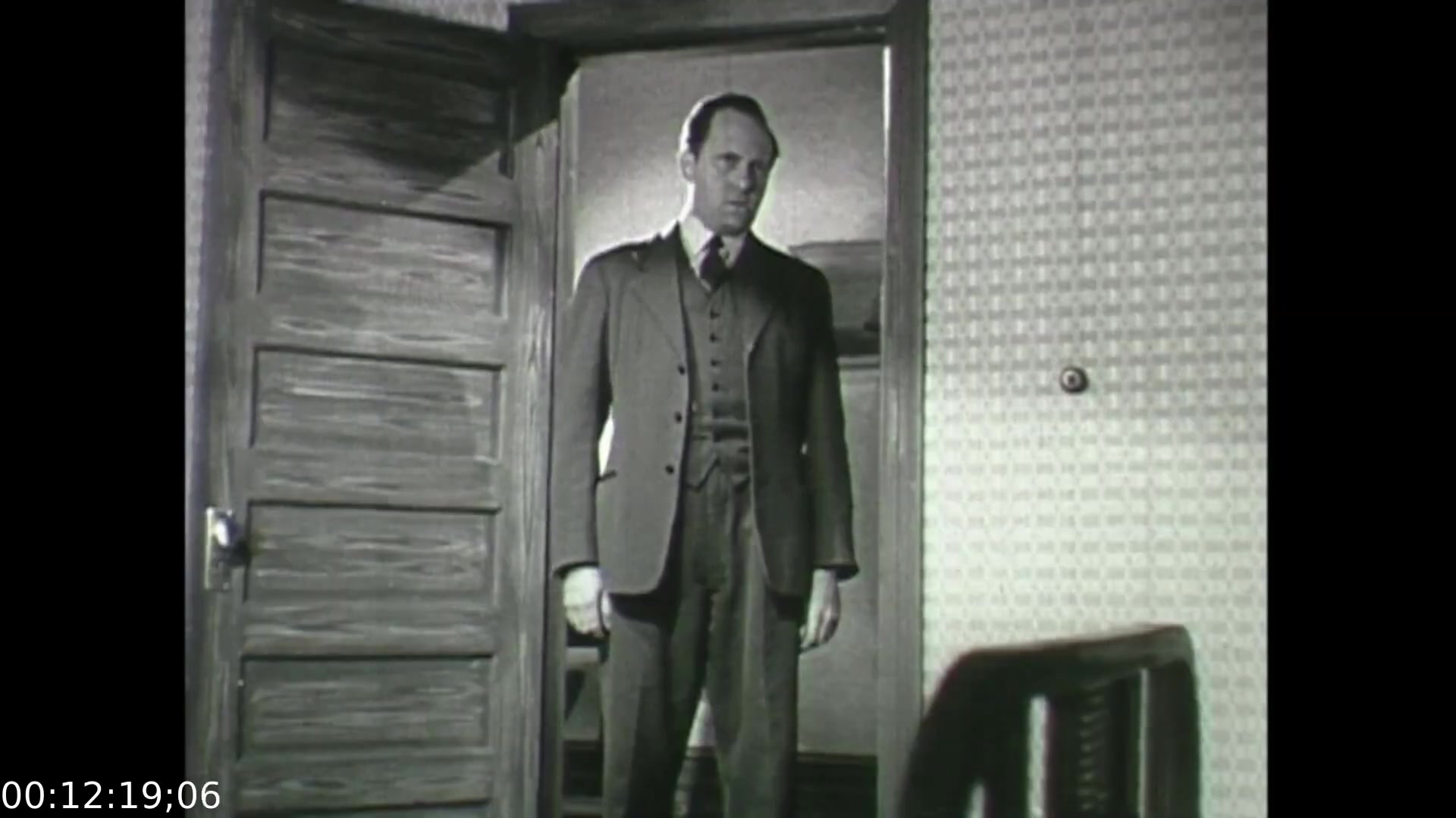 The still from 'Feelings of Depression' (1950) captures a somber moment featuring John Murray, a businessman experiencing emotional turmoil. He stands in a doorway, possibly symbolizing a transition between his internal struggle and the external world. His formal attire reflects his role as a conscientious individual, while his expression hints at despondency and the weight of his psychological challenges. The setting, with its simple decor, underscores his isolation, a theme central to the film’s exploration of depression and mental health awareness.