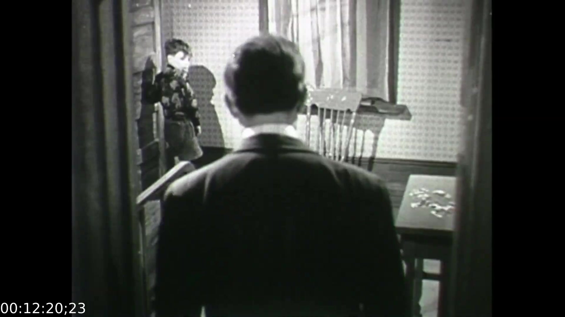 The still from 'Feelings of Depression' depicts a poignant moment reflecting the emotional struggle of the protagonist, John Murray. In the image, a man in a suit stands with his back to the camera, suggesting distance or isolation as he faces a table with scattered items, possibly symbolizing disarray in his life. A child in a patterned sweater stands in the background, potentially representing innocence or the emotional ties that complicate the adult's despondency. The setting, characterized by a modest interior, underscores the personal and societal context of mental health issues in the 1950s. This educational dramatization aims to foster understanding of depression's impact on both the individual and their loved ones.