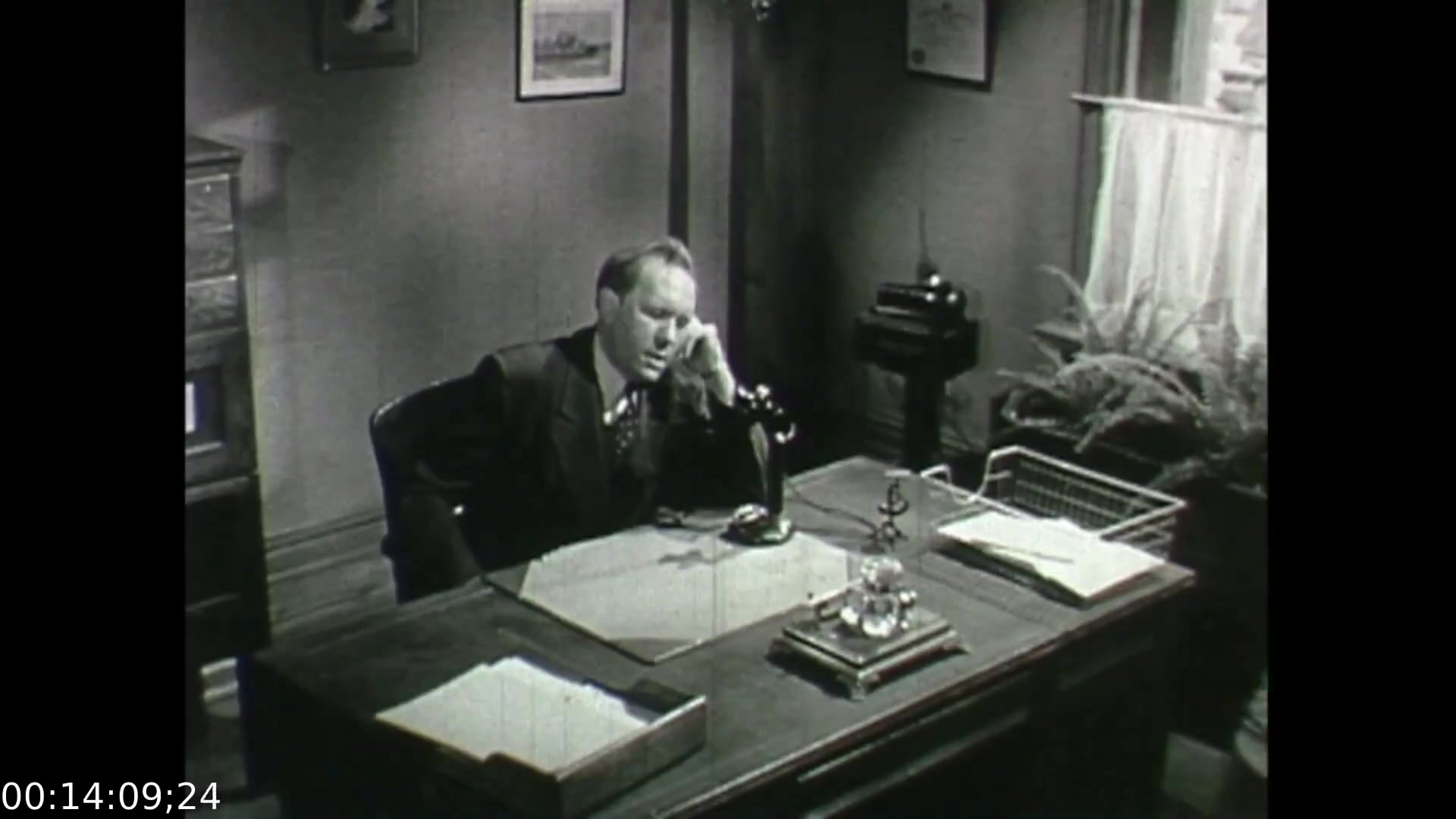 The still from 'Feelings of Depression' (1950) shows a somber office scene featuring a man, presumably John Murray, seated at a desk. He appears to be deep in thought or distress, resting his face on one hand while holding a phone receiver in the other. The decor is simple, with documents and a glass of water on the desk, reflecting a typical mid-20th century office environment. The mood is heavy, capturing the essence of the film's focus on the emotional struggles associated with depression.